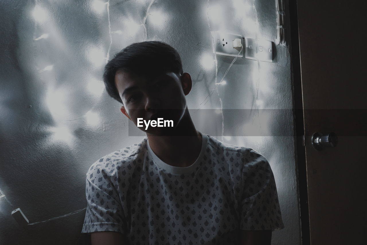 Portrait of young man standing by illuminated string lights on wall in darkroom