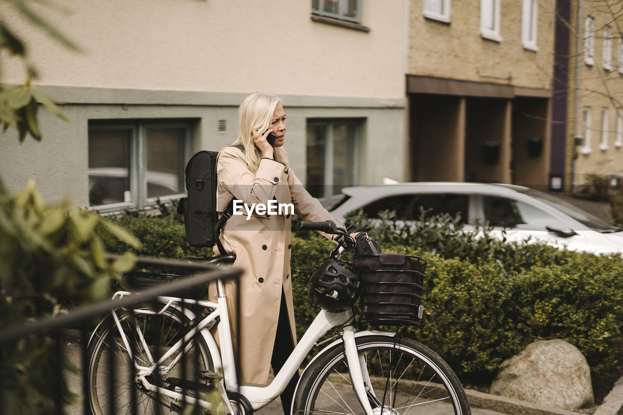 Businesswoman with electric bicycle talking on smart phone while standing outside building