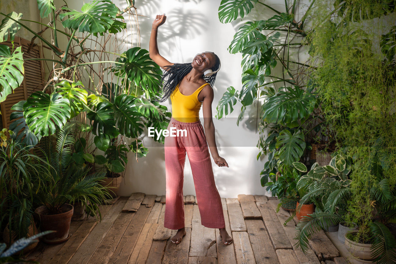 Happy carefree black woman dancing with closed eyes listening music in headphones among houseplants