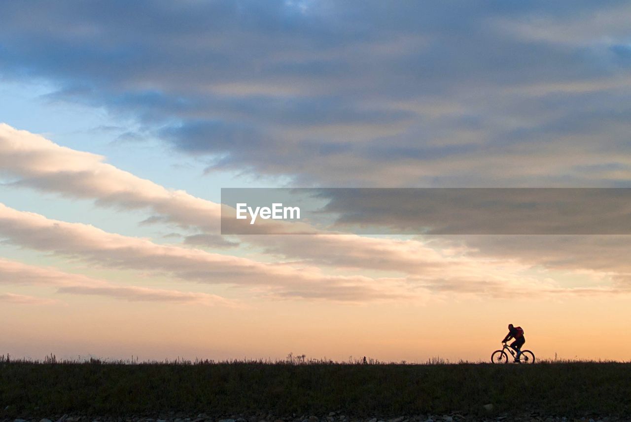 Man riding bicycle on grassy field against cloudy sky during sunset