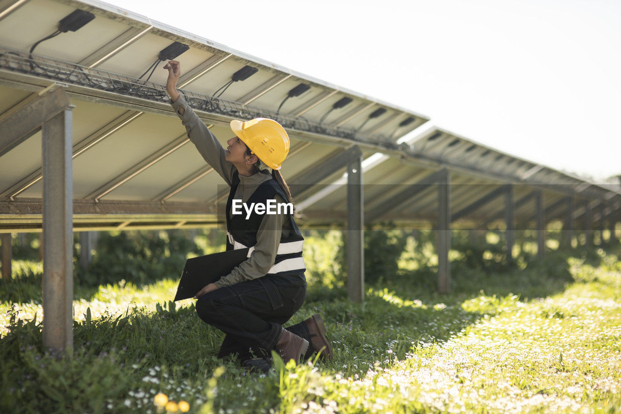 Female engineer fixing connection of solar panels while kneeling at power station