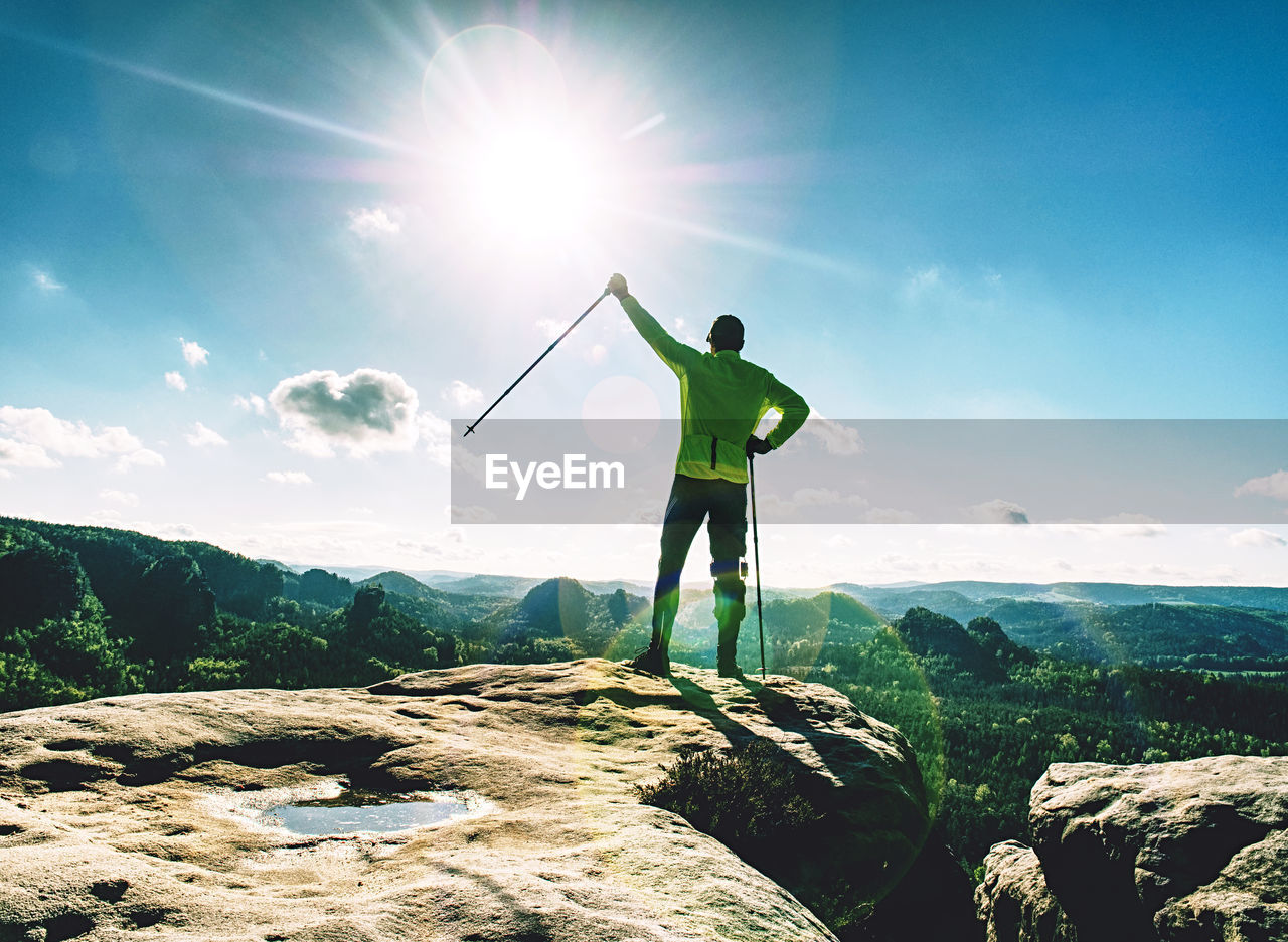 Funny slim blond tourist guy holds trekking stick on rocky top. man in yellow black spoert jersey