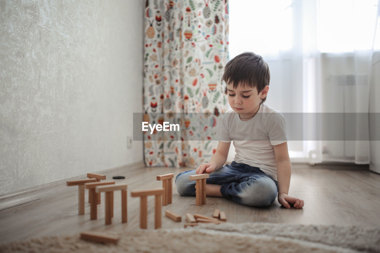 Boy caucasian brunette sad and serious plays on the floor, builds towers from wooden designer, 
