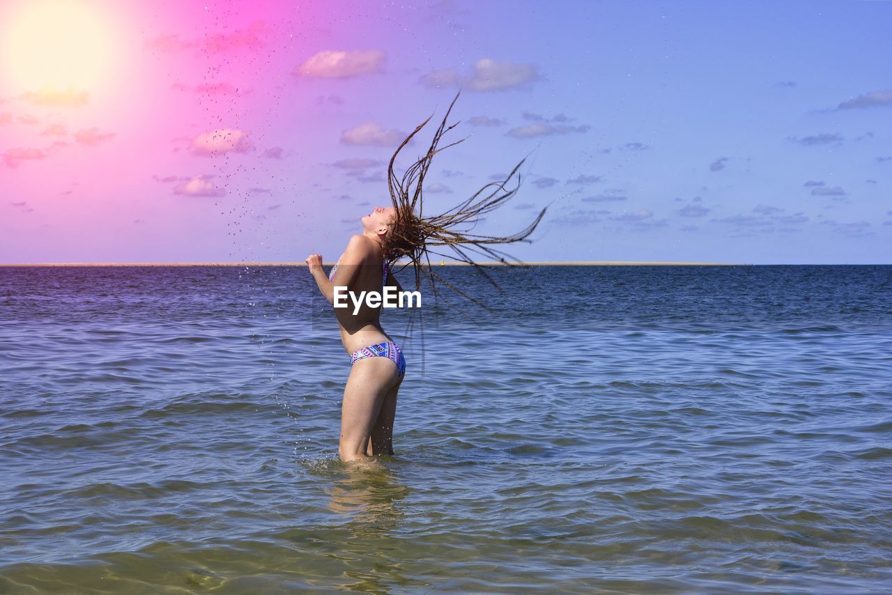 Side view of girl splashing water while tossing hair in sea against sky