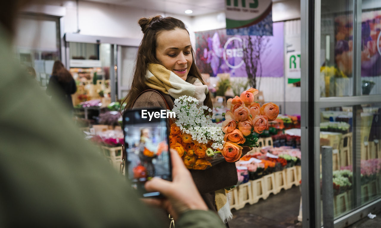 A woman takes a photo with her mobile phone of another woman with a bunch of peonies