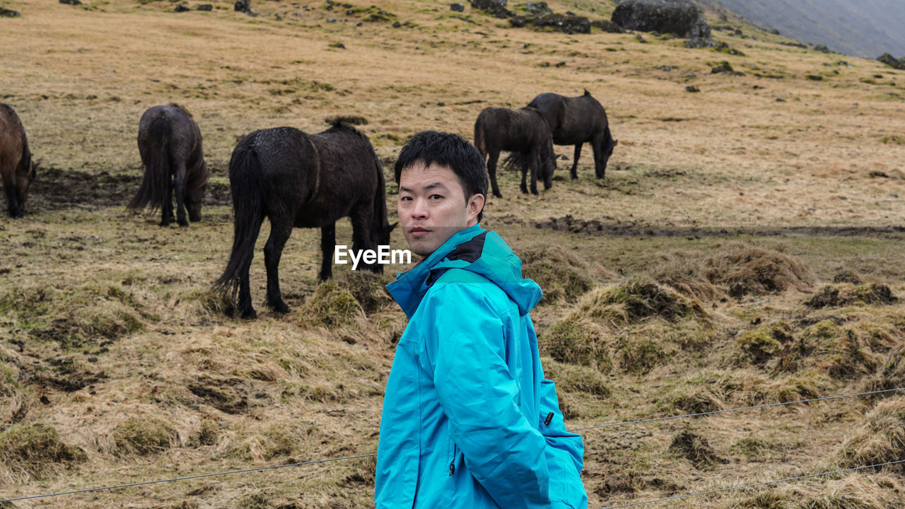 Portrait of man standing against horses grazing on field