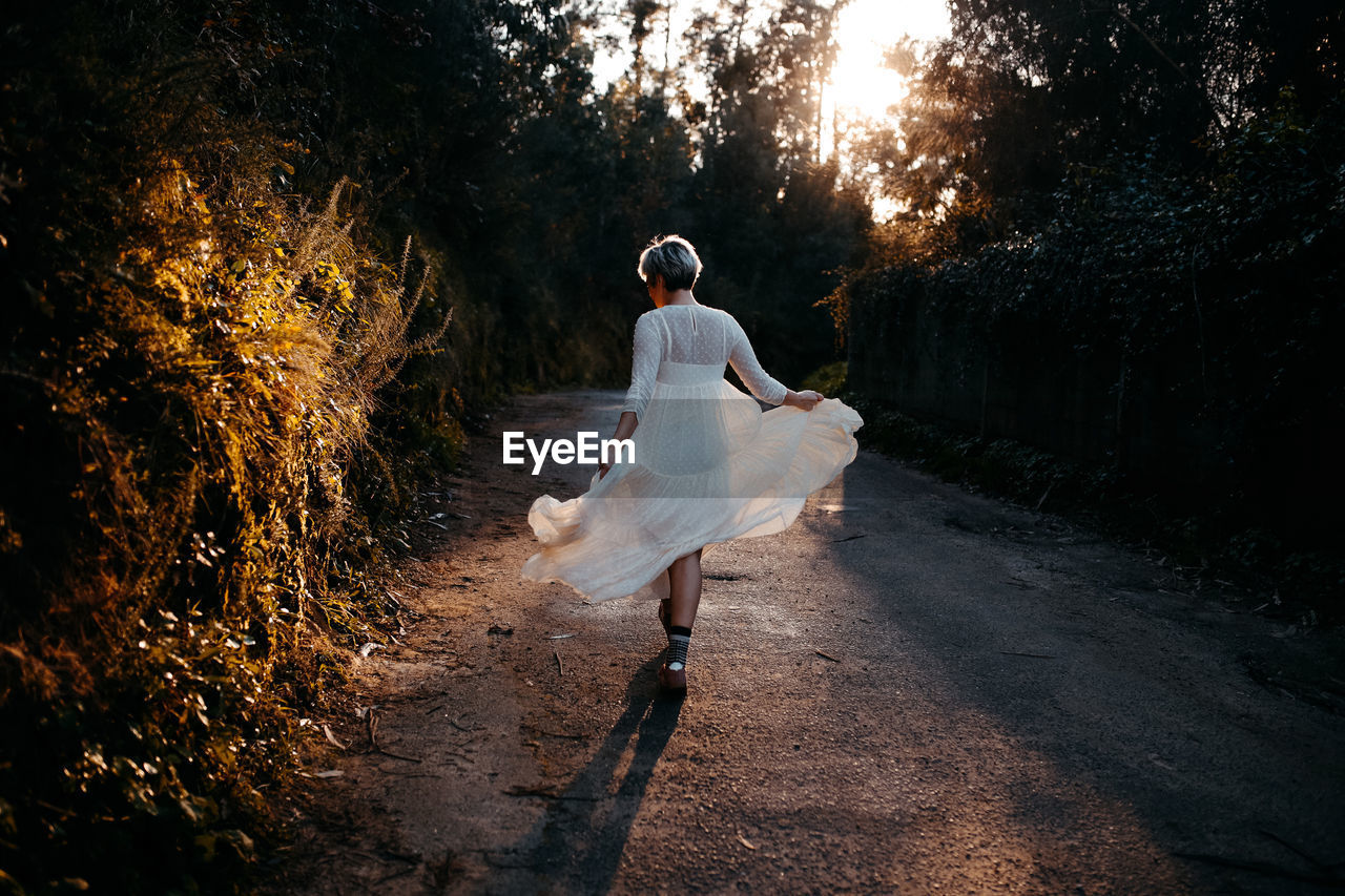 Full body back view of anonymous female wearing white dress walking on rural road among green trees in nature on evening time