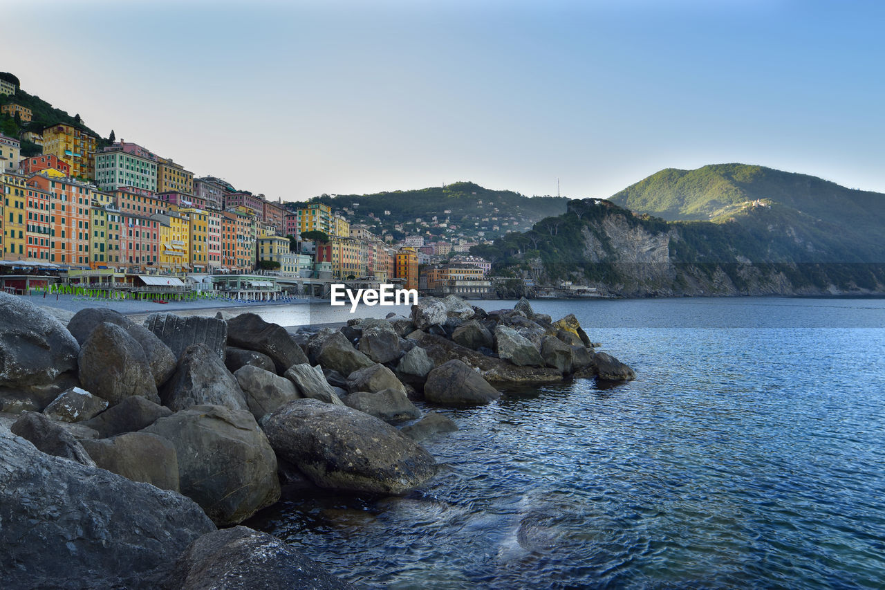 ROCKS IN SEA BY BUILDINGS AGAINST SKY