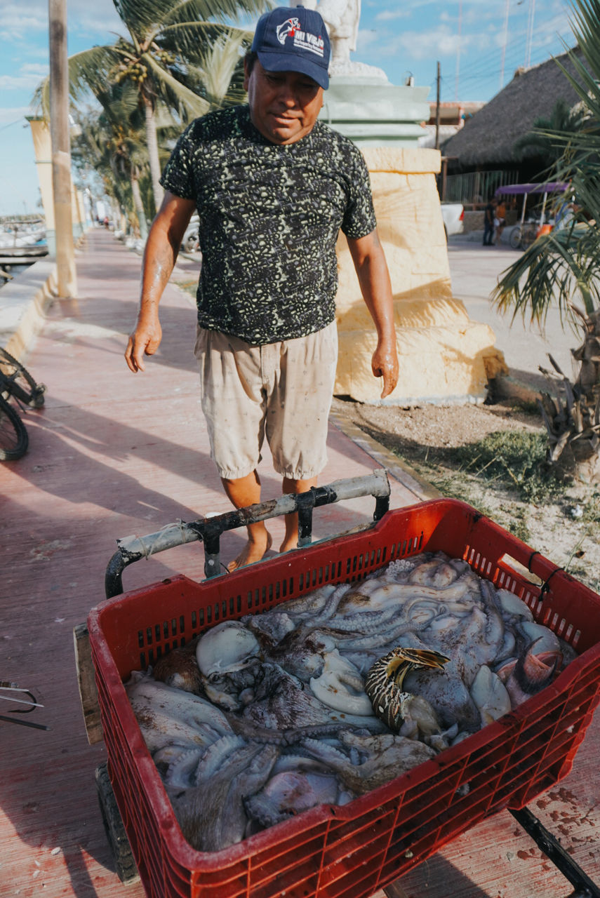 one person, men, day, full length, transportation, nature, adult, standing, fish, occupation, fishing, front view, water, casual clothing, sea, person, outdoors, seafood, mode of transportation, food, hat, fishing industry, clothing, food and drink, nautical vessel, animal, looking at camera, sunglasses, vehicle, beach, holding