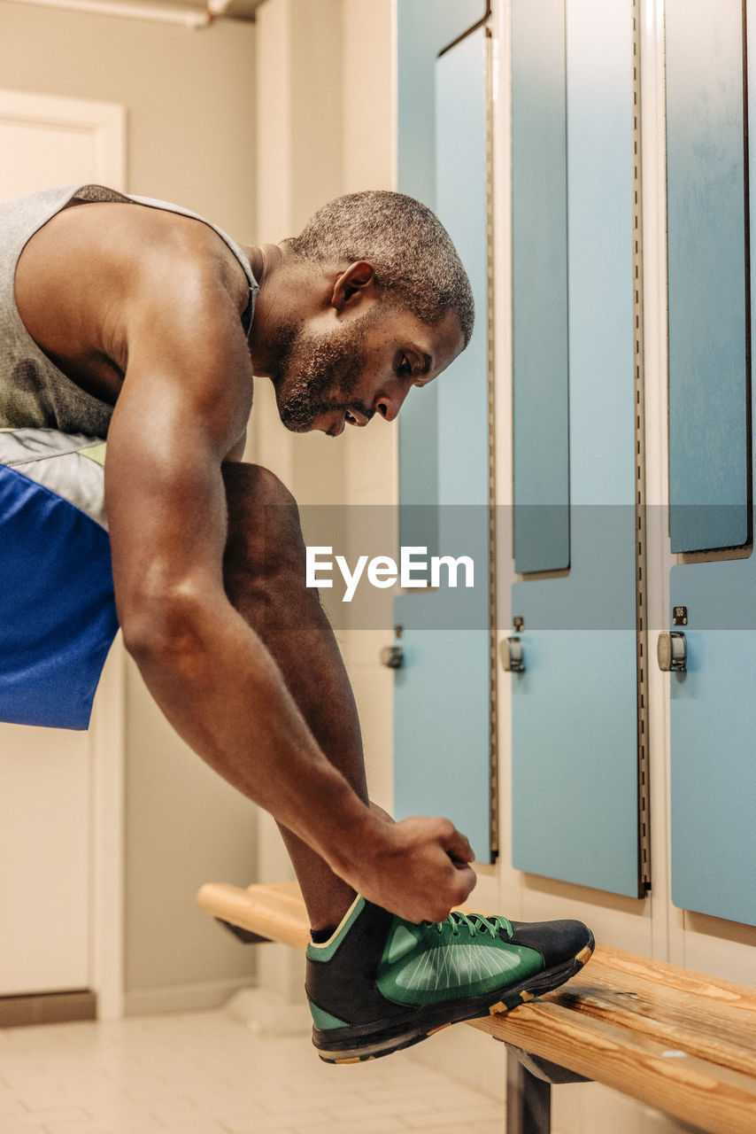 Male athlete tying shoe lace by bench in locker room