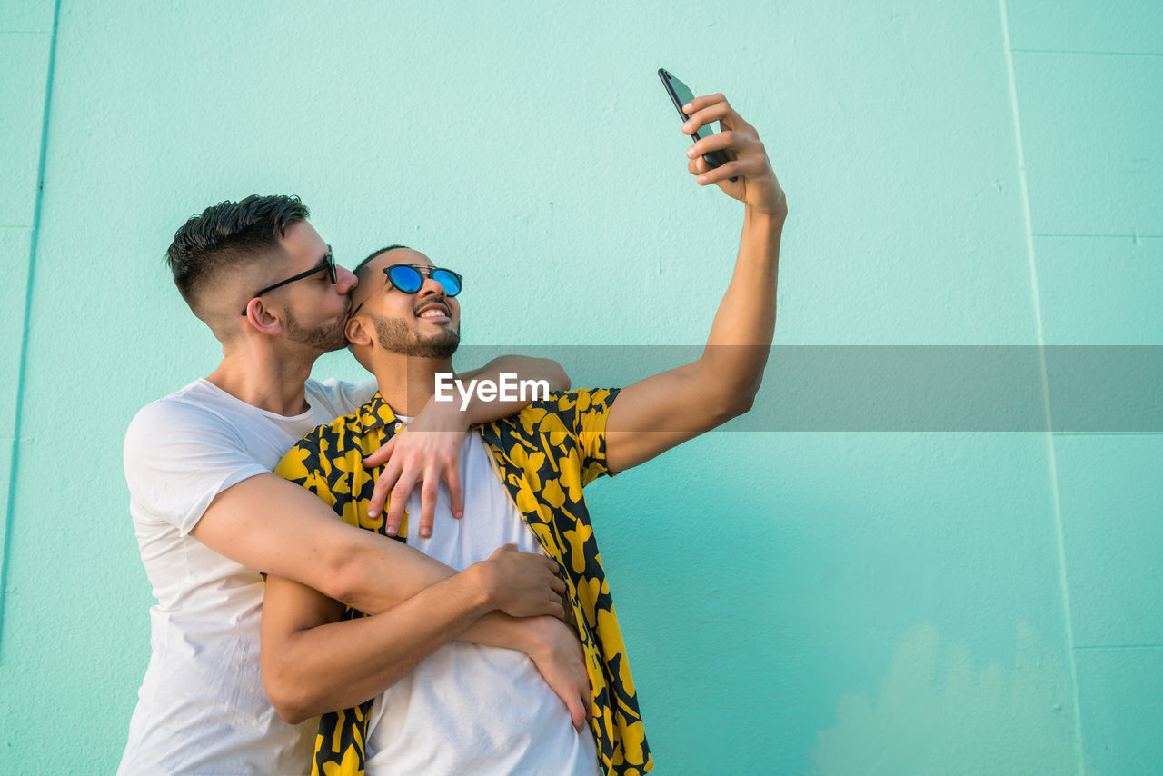 Gay couple taking selfie while standing against wall