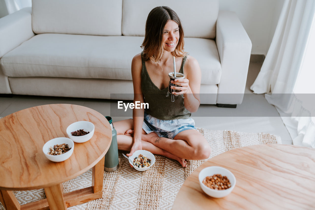 High angle of delighted female with tasty drink and bowl of healthy granola sitting on floor at home and having nourishing breakfast