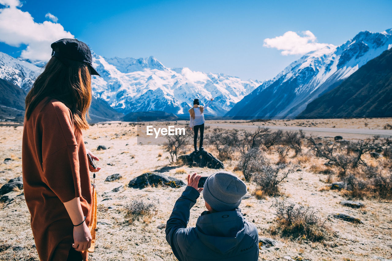 REAR VIEW OF WOMAN PHOTOGRAPHING ON SNOWCAPPED MOUNTAINS