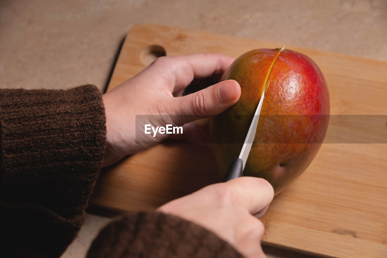 Close-up of woman's hands cutting fresh mango on wooden cutting board at home kitchen