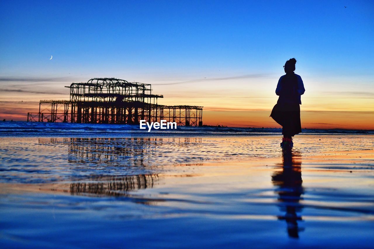Silhouette woman standing on shore while looking at ruined west pier during sunset