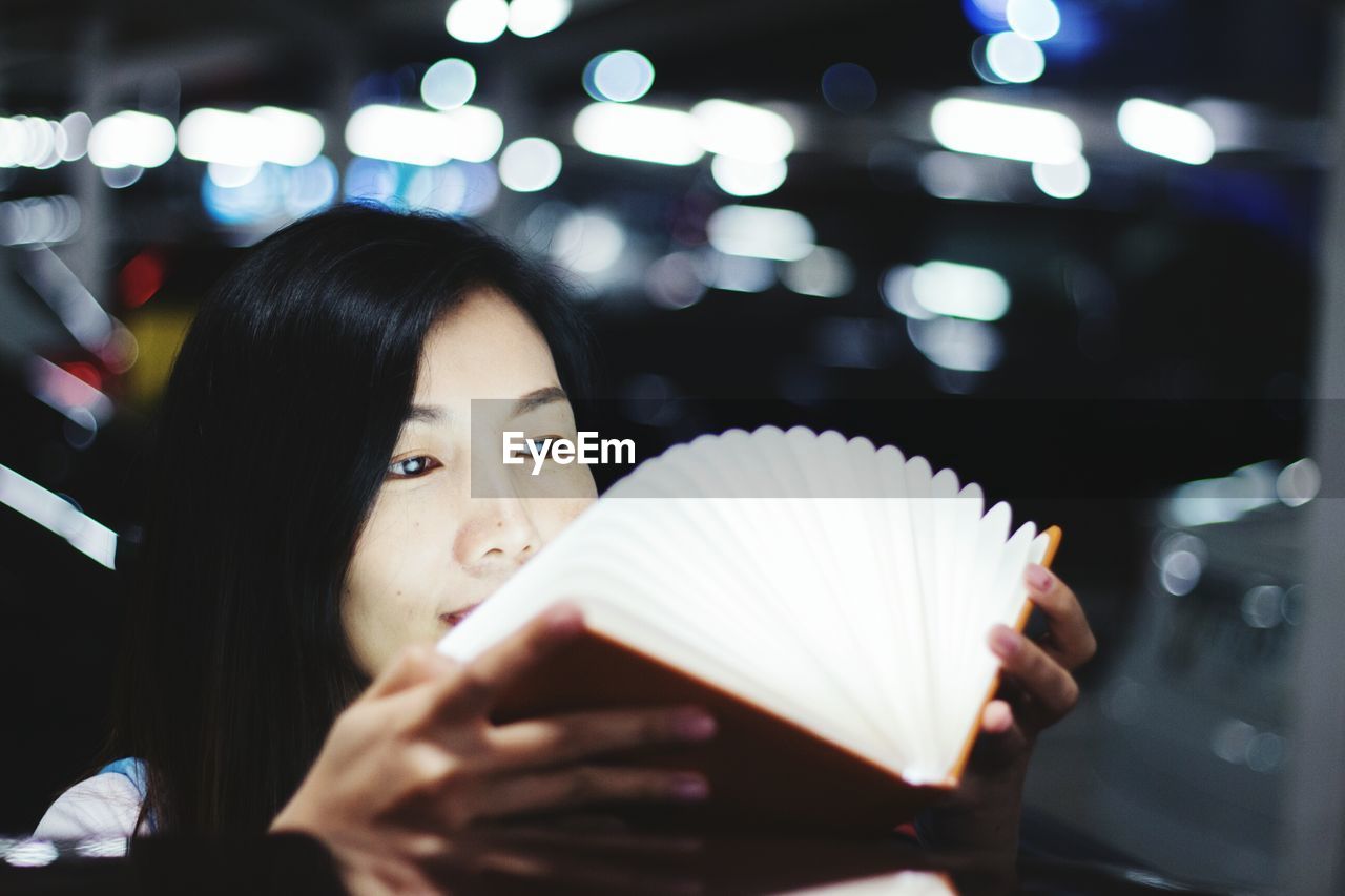 Young woman holding book against illuminated lights at night