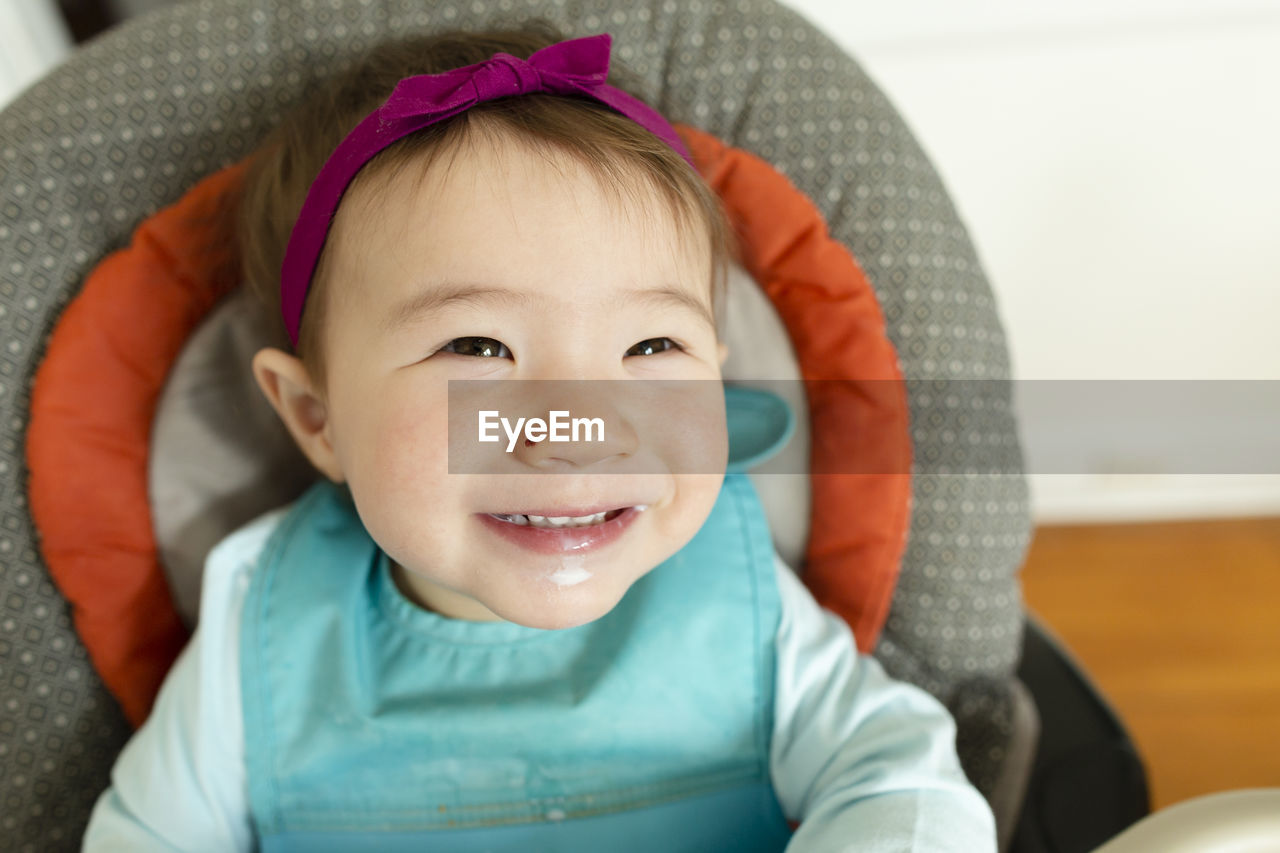 Adorable smiling baby girl wearing headband sits in highchair at home