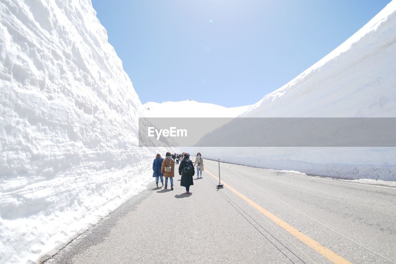 REAR VIEW OF PEOPLE WALKING ON ROAD AMIDST SNOWCAPPED MOUNTAINS