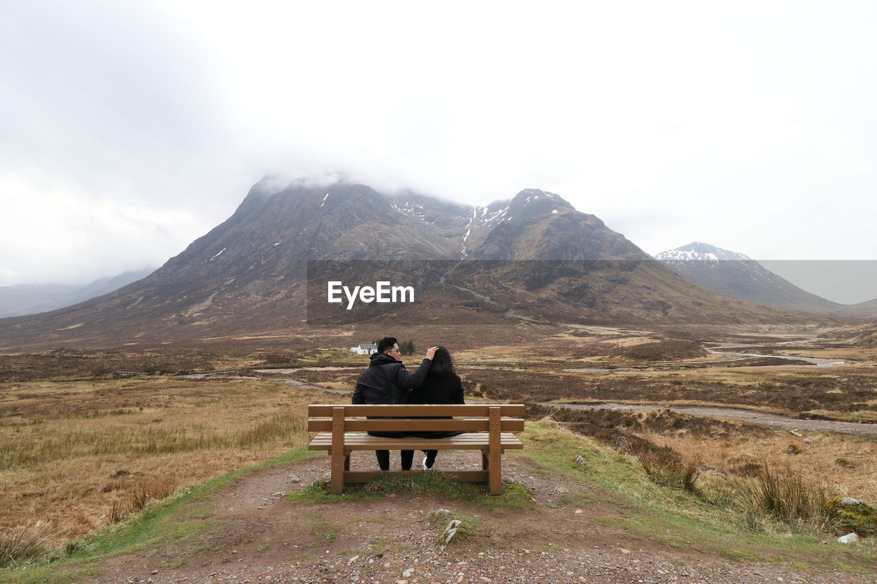 REAR VIEW OF COUPLE SITTING ON BENCH AGAINST MOUNTAIN