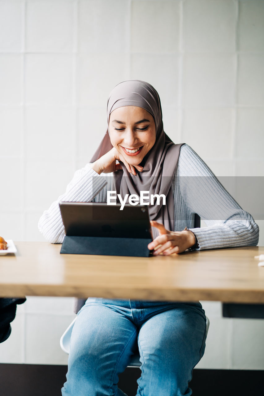 Content muslim female in hijab and talking on video chat via tablet while sitting at table in cafe