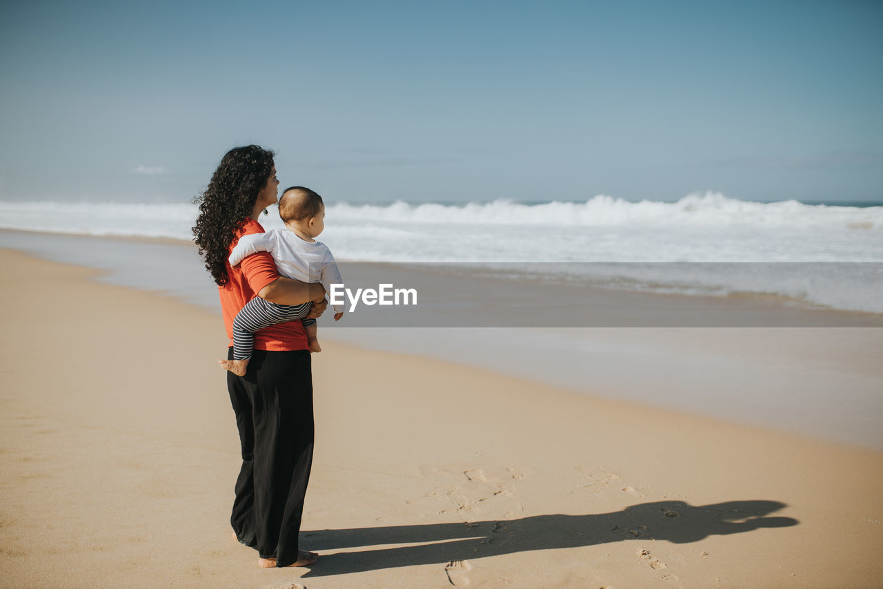 Full length of mother with sun standing on beach against sky