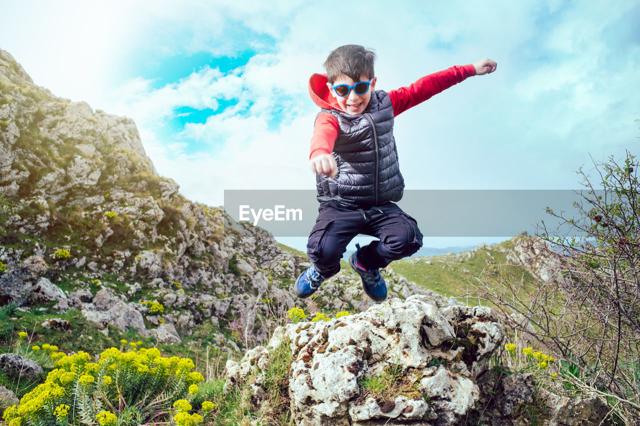 Smiling child during a mountain trek jumps from a rock amused under a blue sky
