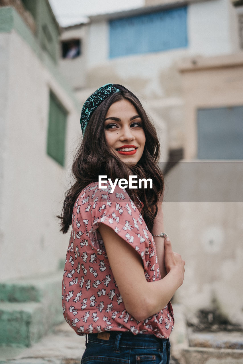 PORTRAIT OF SMILING WOMAN STANDING AGAINST WALL