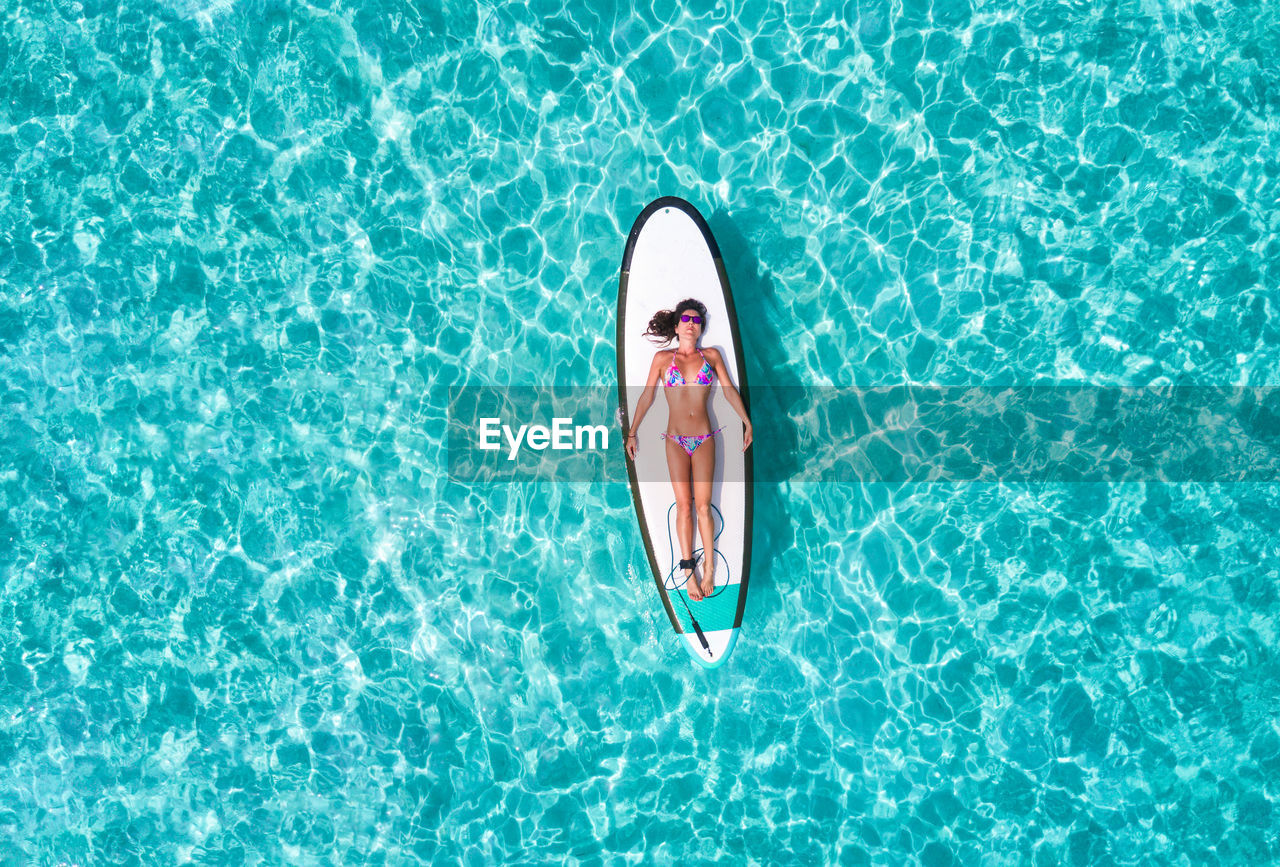 Aerial view of woman in bikini lying on surfboard over sea