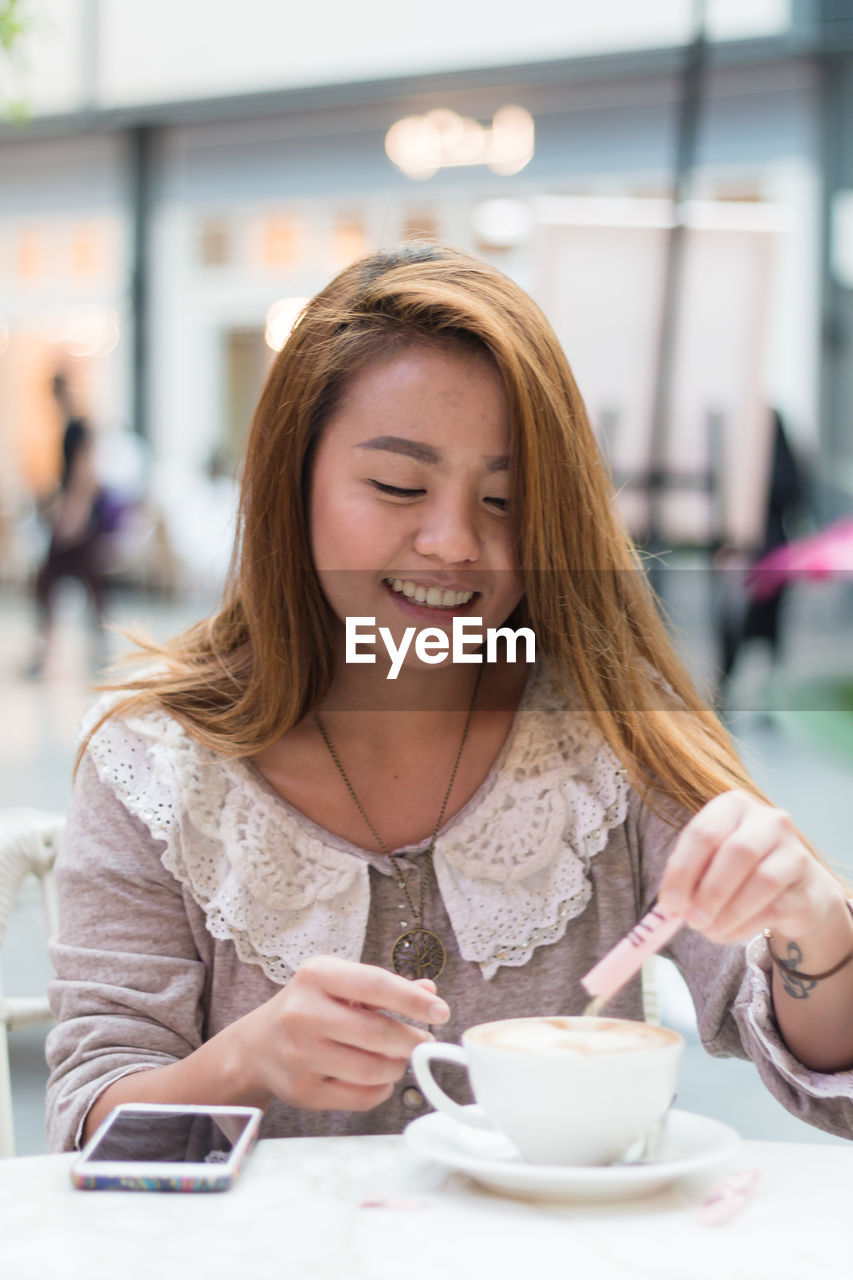 CLOSE-UP OF SMILING WOMAN HOLDING COFFEE WHILE SITTING AT CAFE