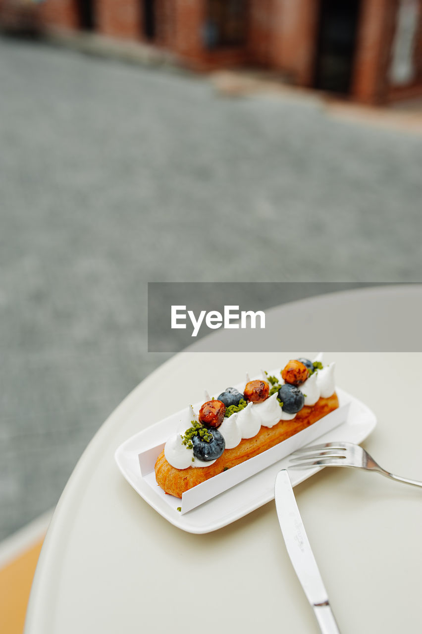 Eclair with white cream, fresh blueberries and caramel in  plate with fork, spoon. outdoor cafe.