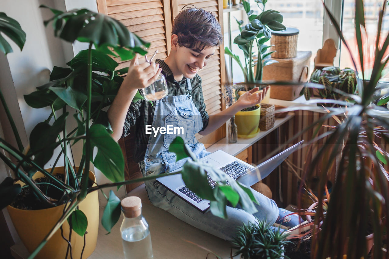 Happy young girl with laptop among plants on balcony, green environment in room, home gardening