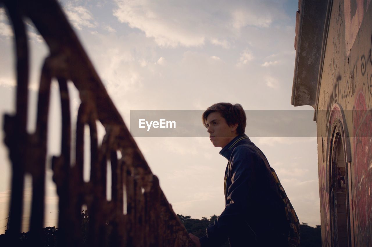 Low angle view of young man standing by railing against sky