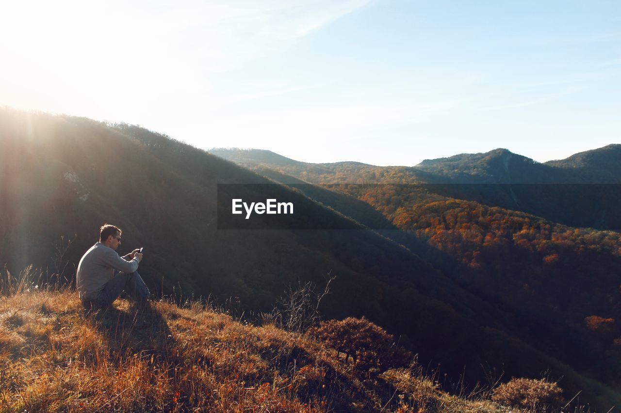 Man using mobile phone sitting on mountain against sky