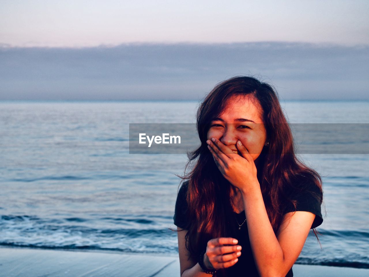 Portrait of beautiful young woman laughing at beach against sky during sunset