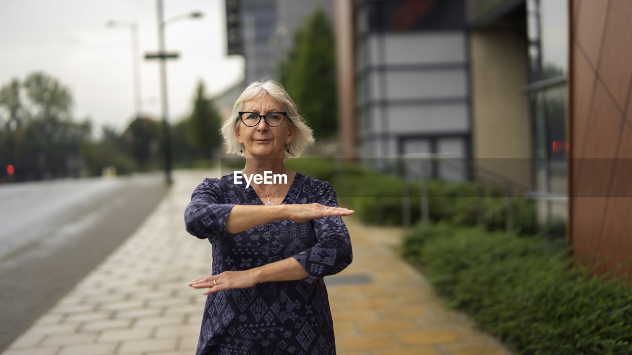 Portrait of woman gesturing while standing on footpath against buildings