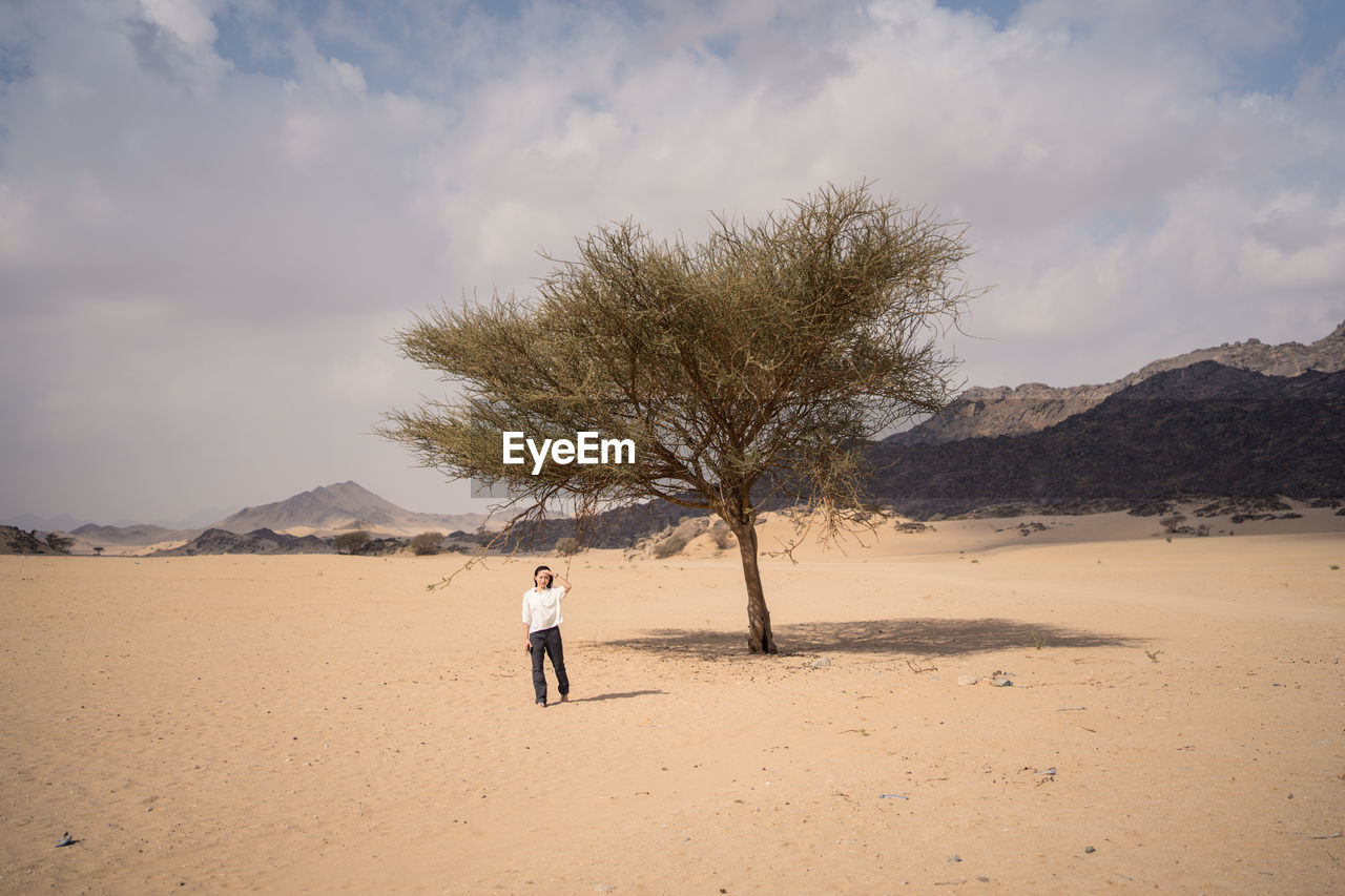 Youthful asian woman looking at camera while standing in middle of sandy desert near a tree in saudi arabia
