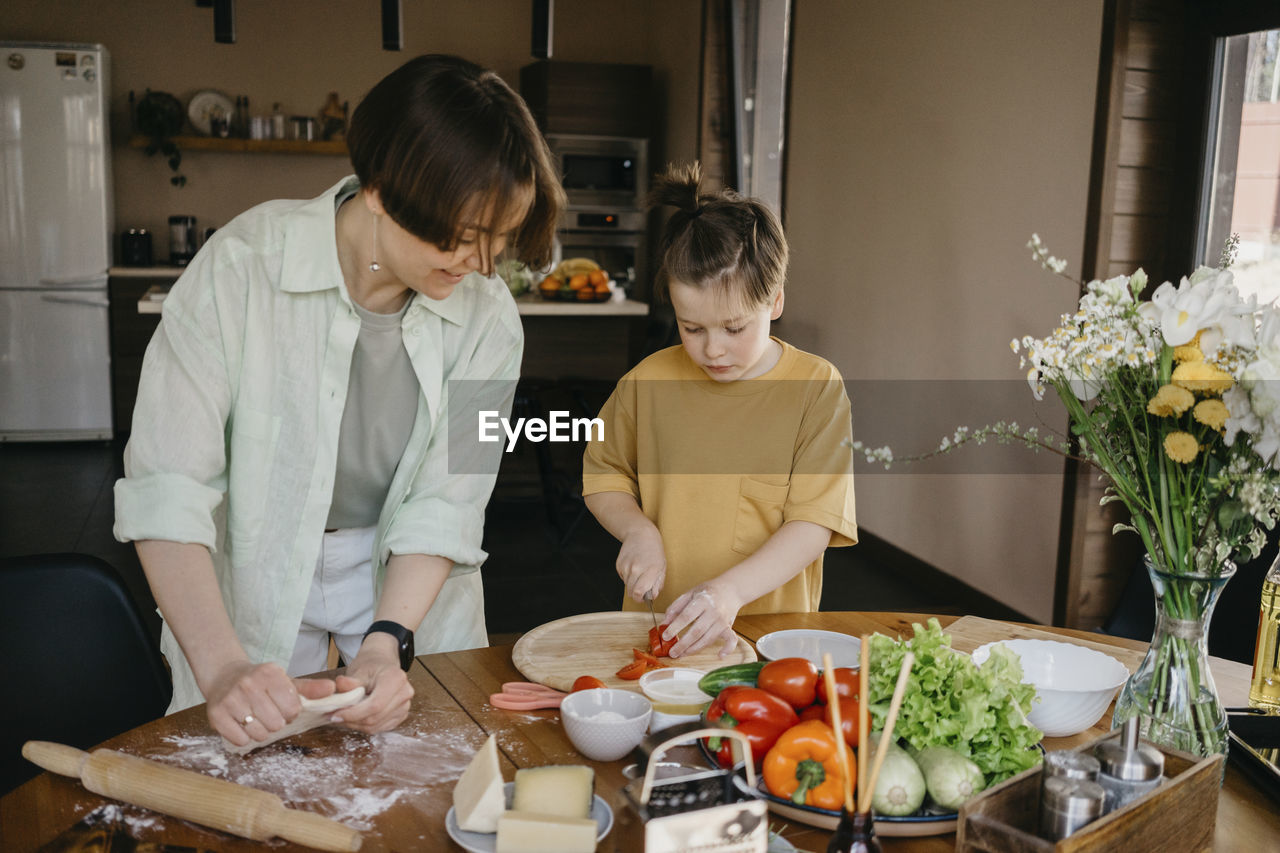 Smiling mother looking at son cutting tomato on table