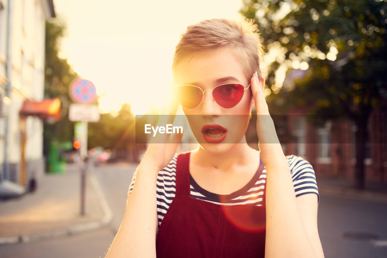 PORTRAIT OF YOUNG WOMAN WEARING SUNGLASSES STANDING ON CITY STREET