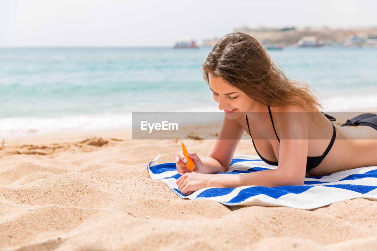 Smiling woman applying sunscreen while lying on towel at beach