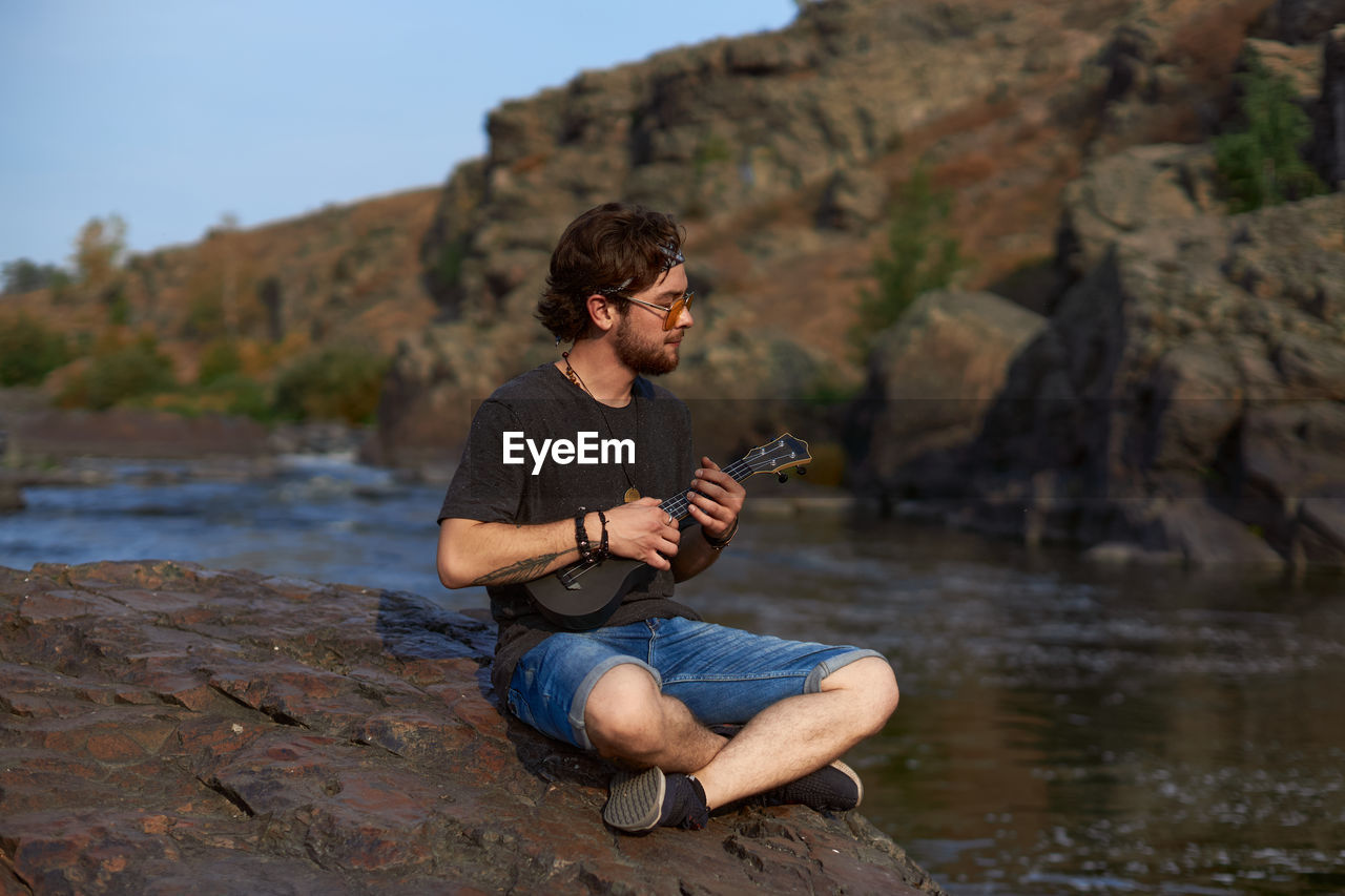 YOUNG MAN USING MOBILE PHONE WHILE STANDING ON ROCK