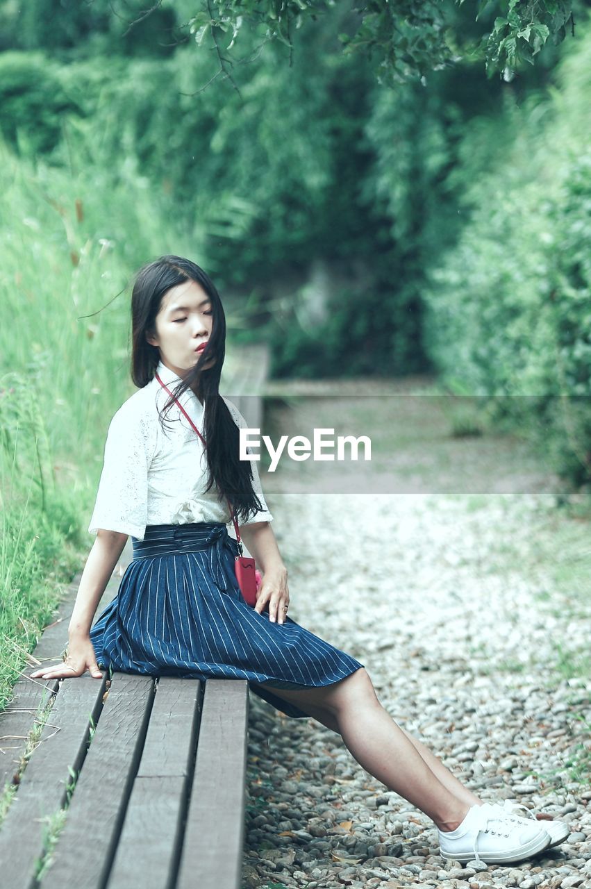 Side view of thoughtful young woman sitting on bench against plants