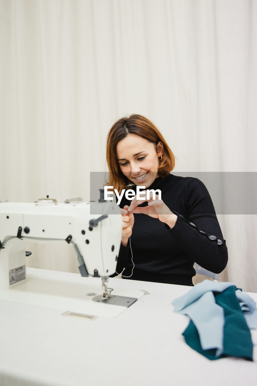 Adult woman sitting at table and making garment part on sewing machine while working in professional studio