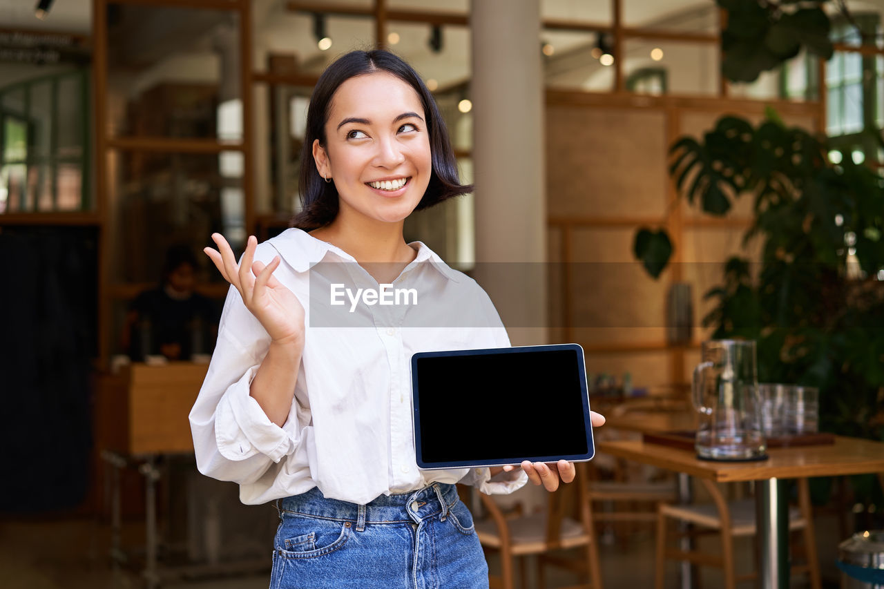 portrait of smiling young woman using digital tablet while standing in cafe