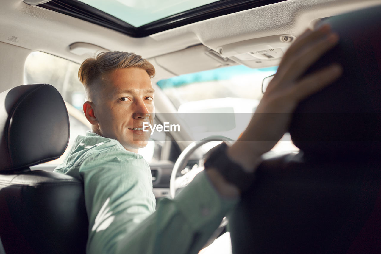 Side view of handsome smiling male driver sitting in parked luxury car and looking at camera