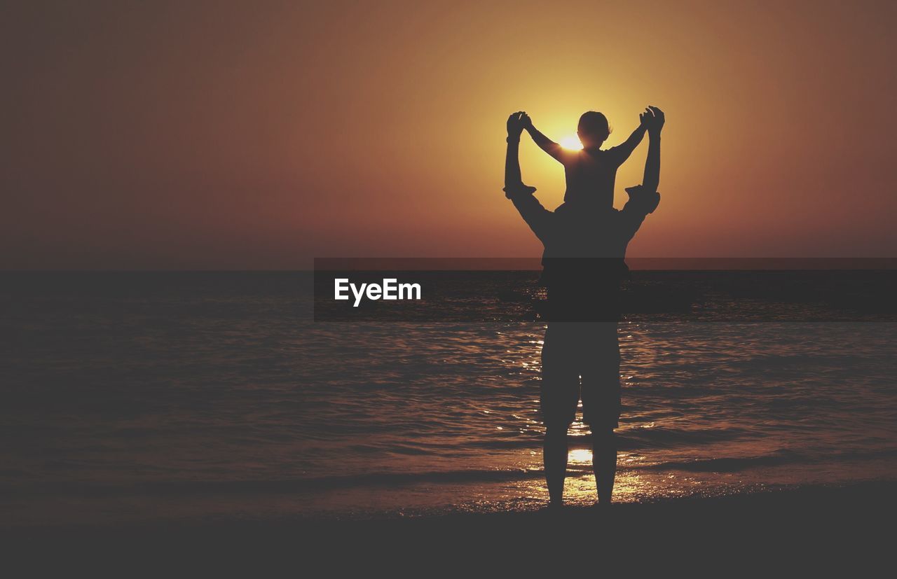 Silhouette man carrying child on beach against sunset sky