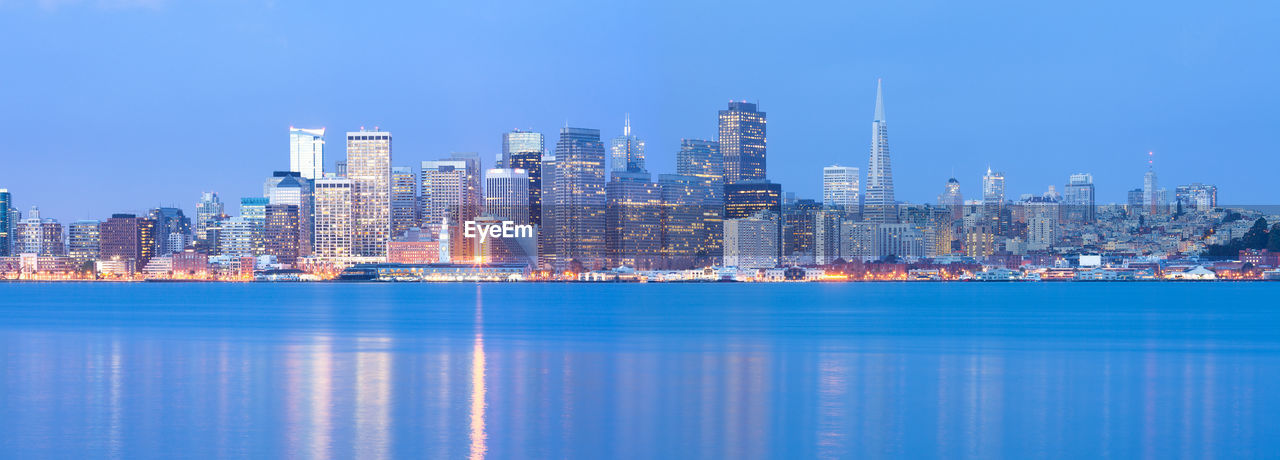 Illuminated modern buildings by bay against blue sky