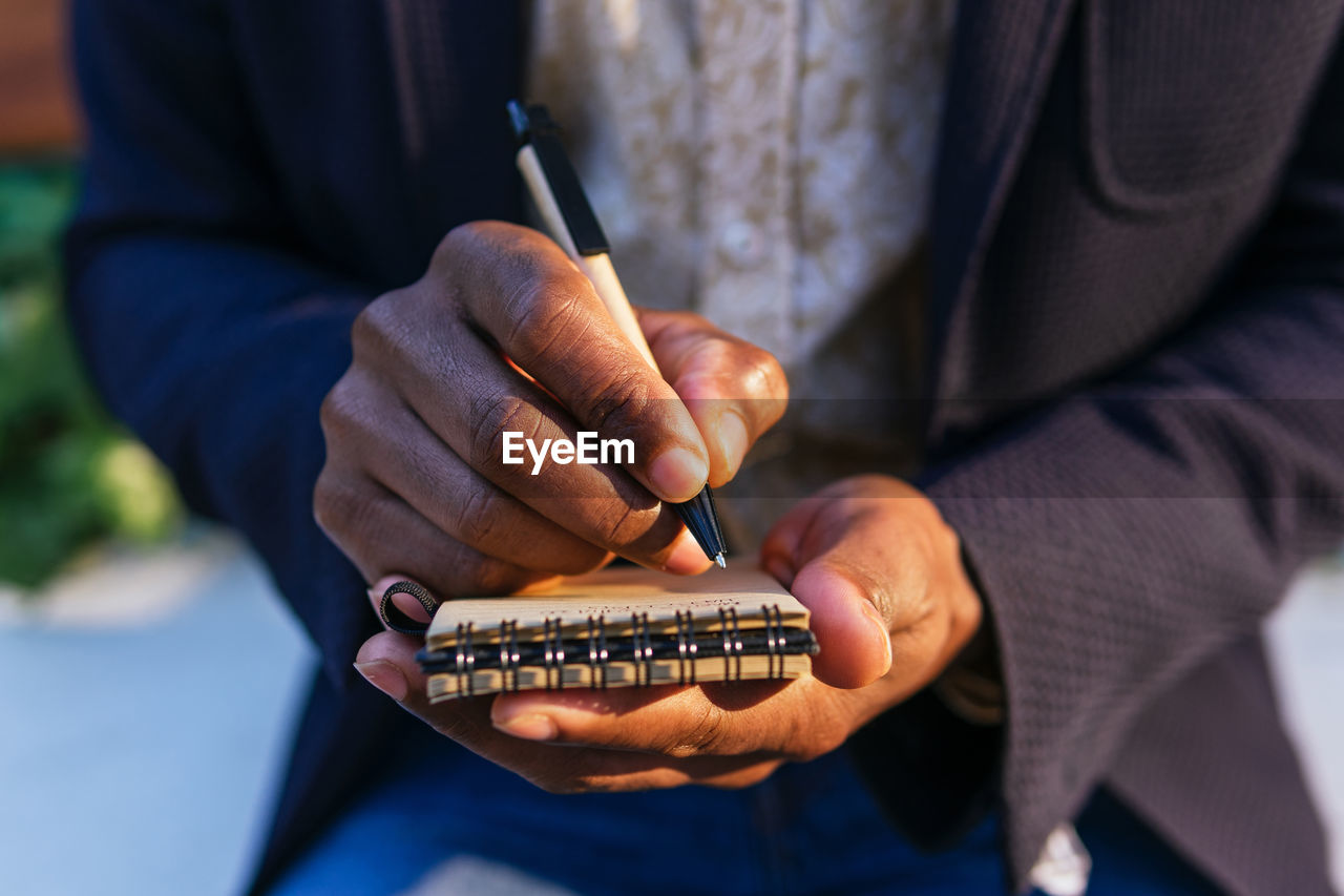 Cropped unrecognizable african american male entrepreneur sitting on bench in urban park and writing plans in notebook