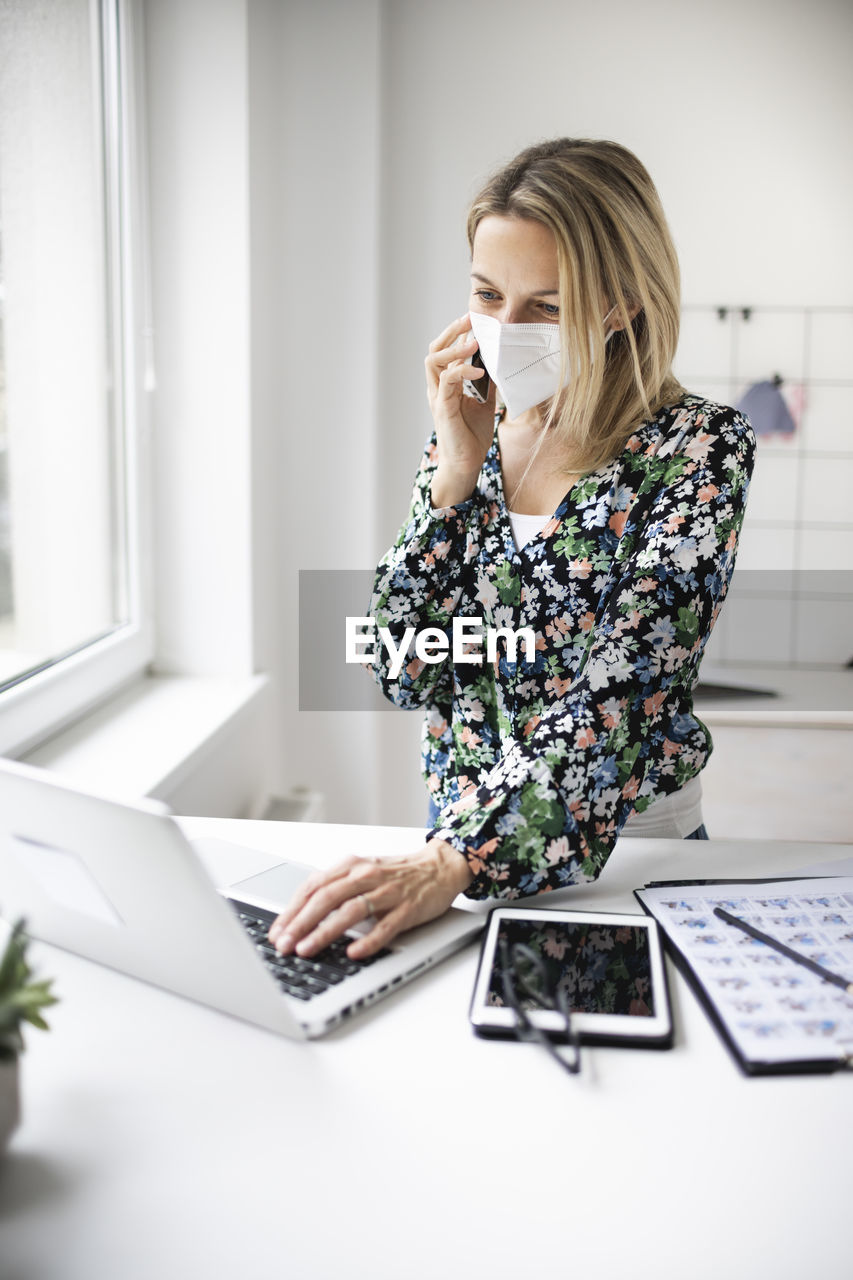 Businesswoman wearing mask using laptop while talking on phone at home