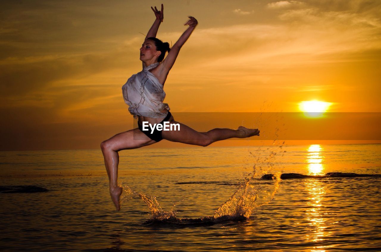 Beautiful woman performing gymnastics in sea against orange sky during sunset