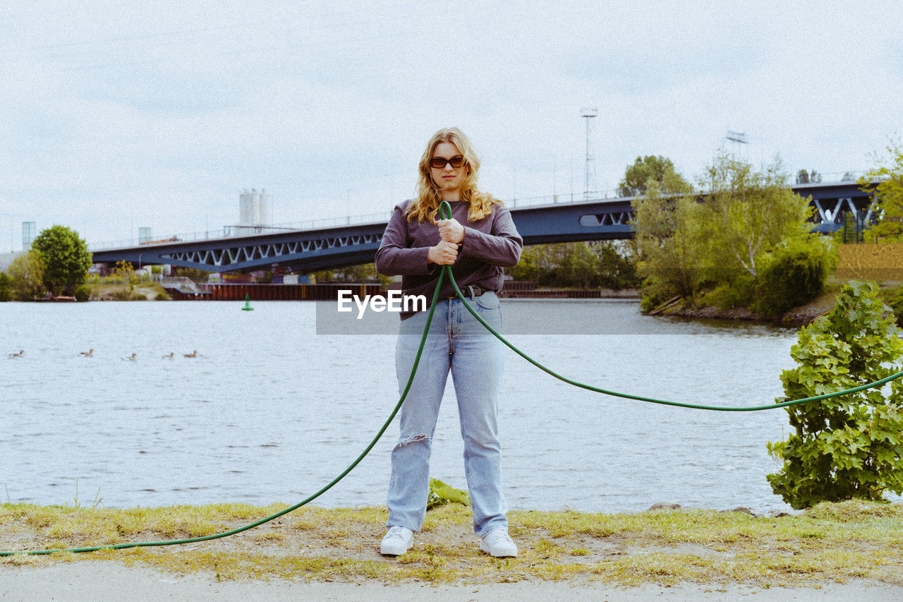 Full length portrait of confident young blond woman holding garden hose
